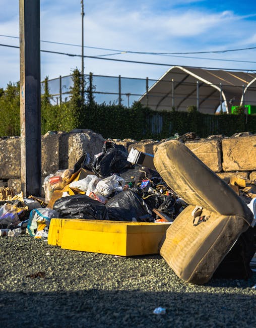 A pile of mixed waste debris and rubbish bags located outdoors on a paved surface, with discarded items including a dirty, aged car tire leaning against the pile. The debris consists of black and clea